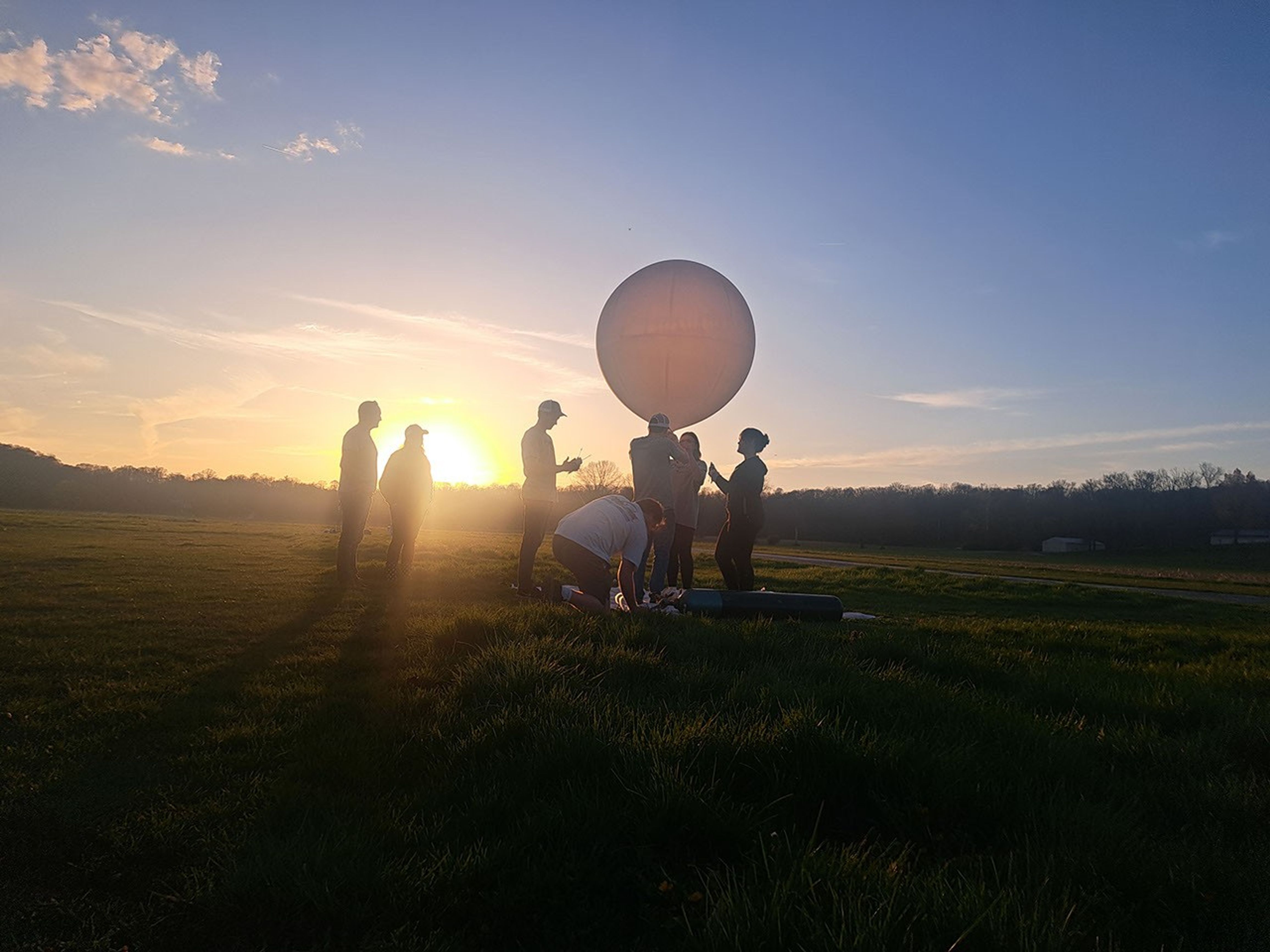 A photograph shows several people in silhouette standing in a large field in front of the setting Sun, gathered around a large, round, white balloon floating just above their heads against a blue sky.